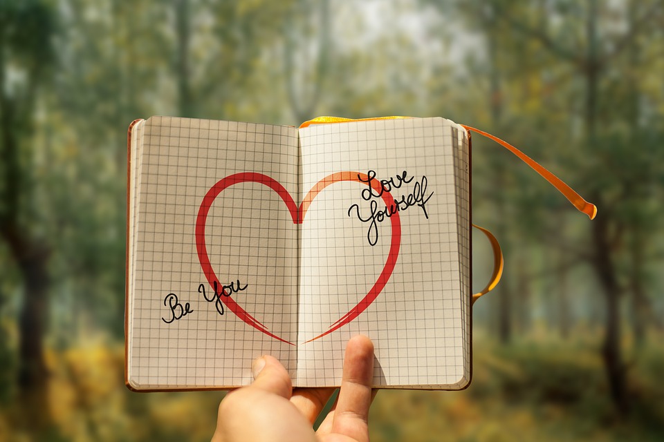 A hand holds open a notebook with a red heart drawn across both pages, with the words 'Be Kind' written inside the heart on the left and 'Stay Sweet' on the right. The notebook is held up against a soft-focus natural backdrop, suggesting a moment of introspection or romantic sentiment.